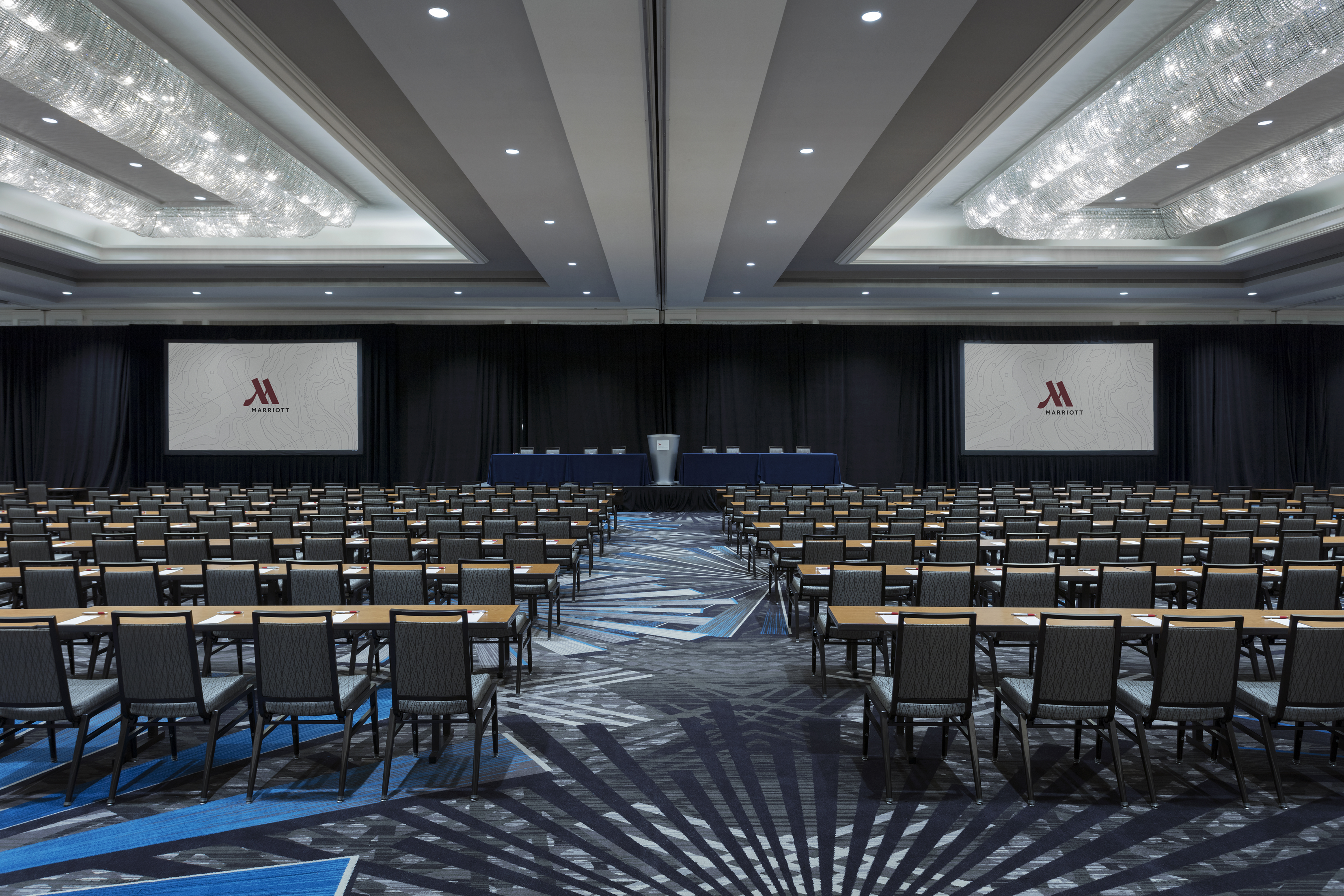 Carpeted ballroom with long rows of tables and chairs facing stage with podium
